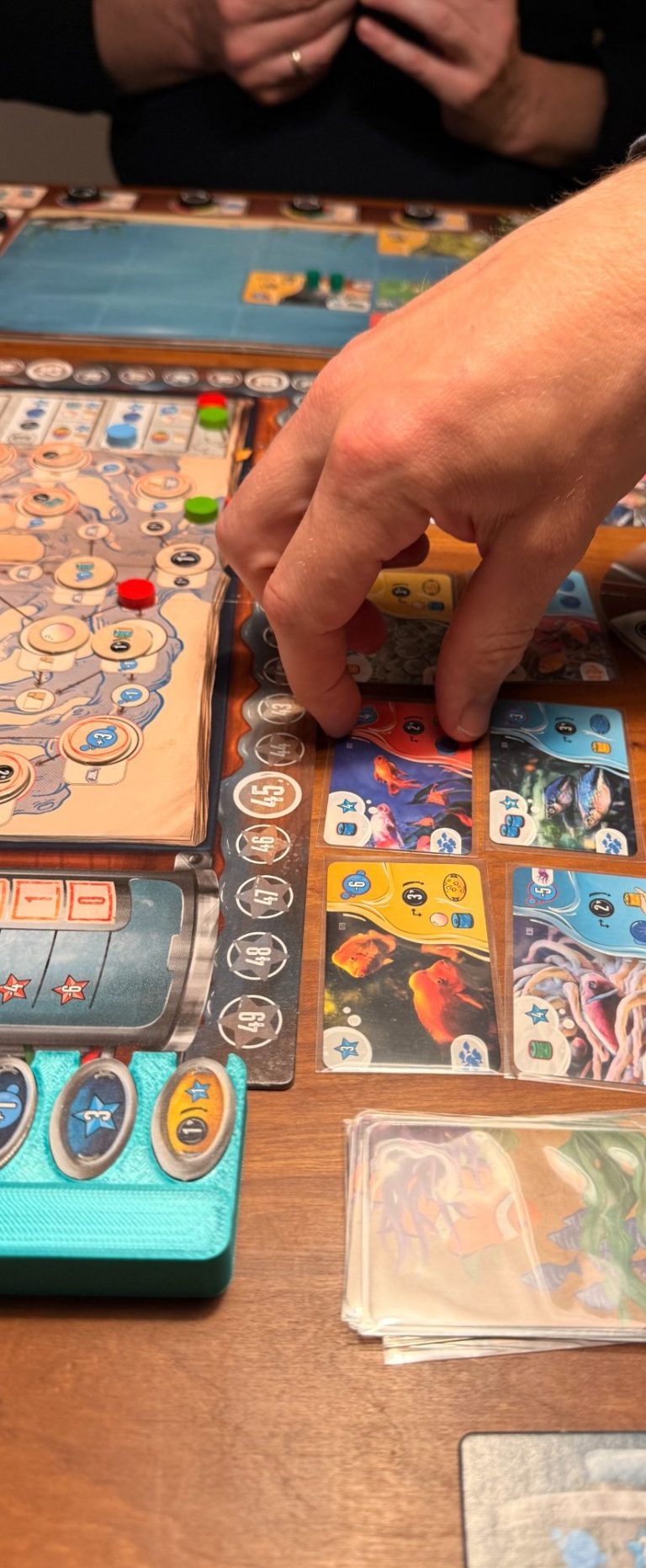 Deckbuilding bei Aquatica. Spieler hält zwei Aktionskarten mit Characteren und Spielbrett im HIntergrund