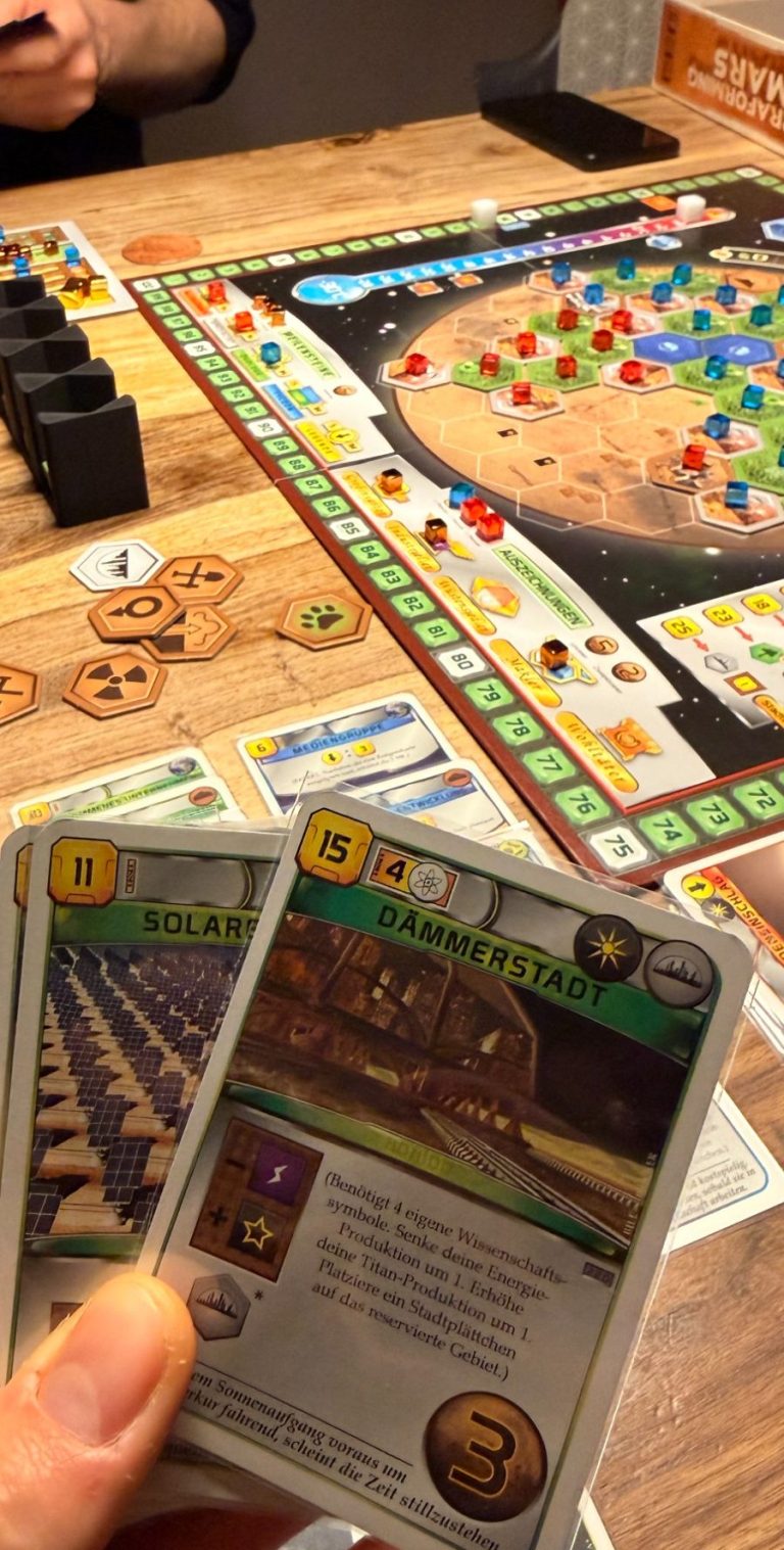 Spieler hält zwei Aktionskarten mit Obst, Nüssen und einem Segelboot, umgeben von Spielbrett.