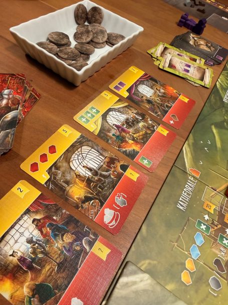 Brettspiel mit Spielkarten, Würfeln und einem Behälter voller Spielsteine auf einem Tisch.