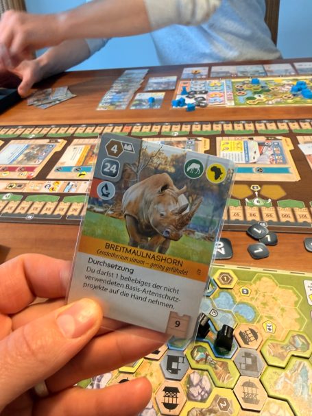 Eine Hand hält eine Spielkarte mit einem Wildschwein, umgeben von Spielmaterialien.