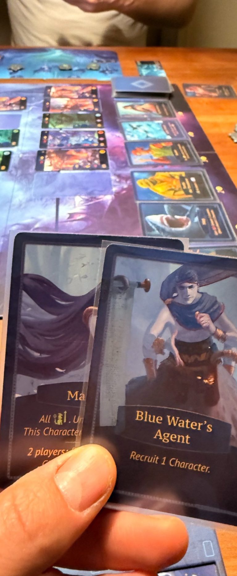 Spieler hält zwei Aktionskarten mit Characteren und Spielbrett im HIntergrund