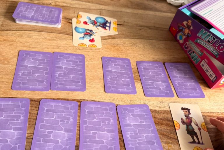 Karten auf einem Tisch, umgeben von Spielmaterialien und einer Spielebox.