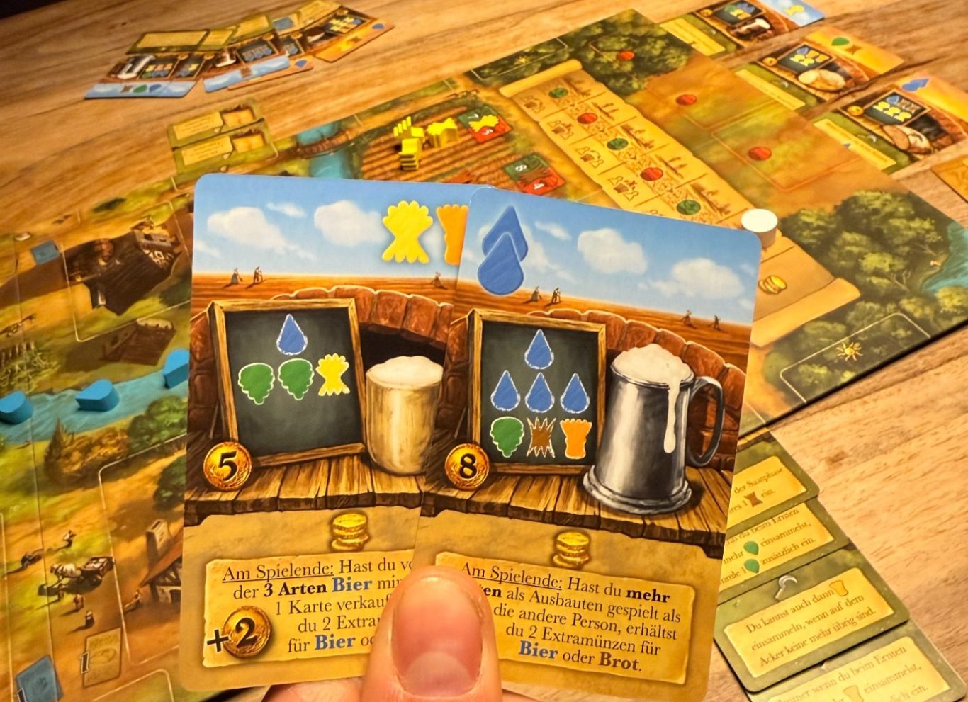 Kartenspiel mit Ressourcenmarkern, Bier und Punktesymbolen auf einem Holztisch.