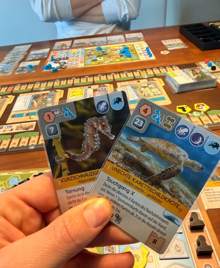 Zwei Spielkarten mit Seepferdchen, auf einem Tisch mit mehreren Brettspielen.