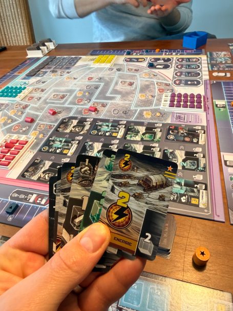 Ein Spieler hält Karten in der Hand, während ein Brettspiel im Hintergrund läuft.