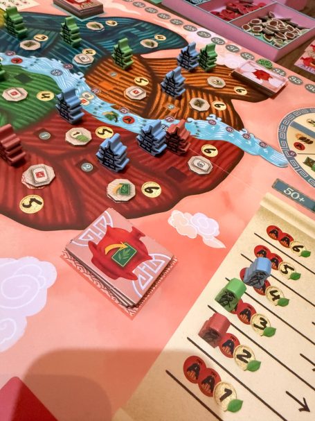 Brettspiel mit bunten Spielfiguren, Ressourcen und einem Fluss auf dem Spielbrett.