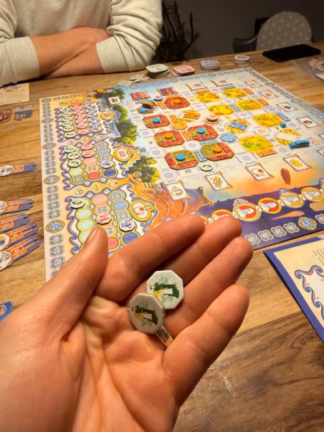 Eine Hand hält zwei Spielmünzen über einem Brettspiel auf einem Tisch.