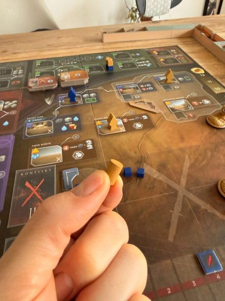 Eine Hand hält eine Spielfigur über einem Brettspiel mit verschiedenen Ressourcen.