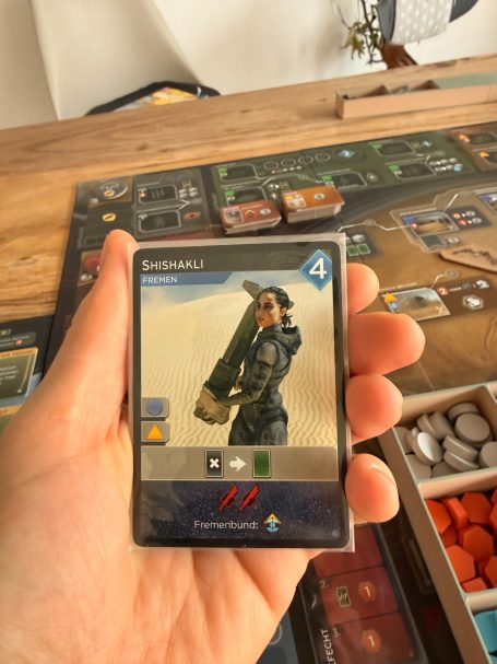 Eine Hand hält eine Spielkarte mit einem Charakter in einer Wüste und einer Waffe.