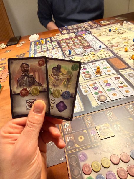 Eine Hand hält zwei Spielkarten über einem Spieleabend mit einem Brettspiel.