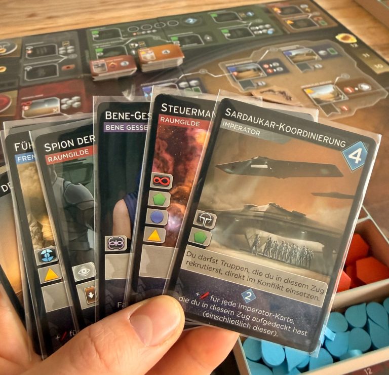 Hand hält fünf Spielkarten vor einem Spielebrett mit bunten Spielsteinen.