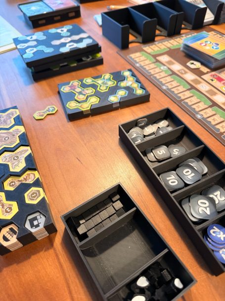 Spielmaterialien auf einem Tisch, darunter Brettspielbrett, Spielchips und Ablageboxen.