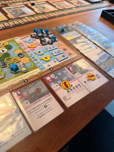 Brettspiel mit verschiedenen Karten, Marker und Spielfeld auf einem Tisch.