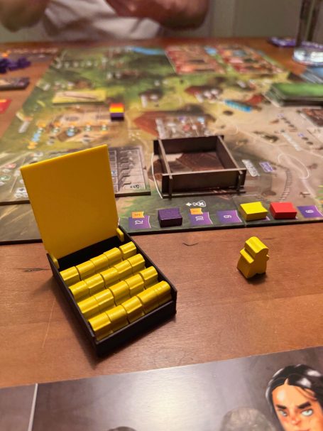 Spielbrett mit gelben Spielsteinen und einem geöffneten Karton auf einem Tisch.